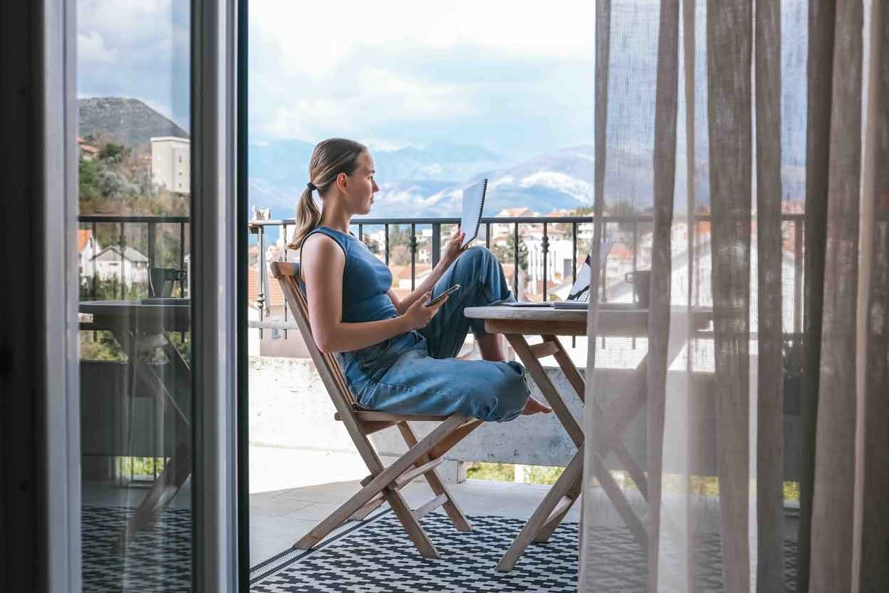 A woman sits on a balcony, using a laptop and smartphone, enjoying a scenic view of mountains and cityscape.